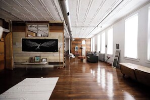 A spacious room with a large flat-screen TV mounted on a wall, wooden flooring, and a long window along one wall.