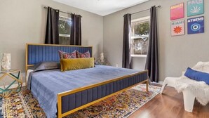 Un dormitorio con una cama, ventilador de techo, ventana con cortinas y cuadros en la pared.