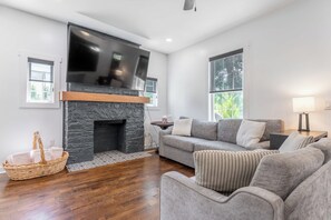 A modern living room with a stone fireplace, a flat-screen TV, a grey sofa, and a wicker basket. - The Northview Nook- Dog-Friendly Nashville Retreat (Nashville)