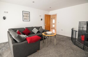 Ein Wohnzimmer mit grauem Sofa, roten und grauen Kissen, einem kleinen runden Tisch und einem schwarzen Metallgeländer.