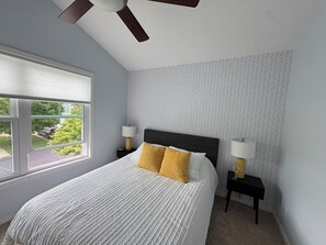 Ein Schlafzimmer mit einem Bett, zwei Nachttischen mit Lampen, einem Deckenventilator und einem Fenster mit Blick auf Grünflächen.
