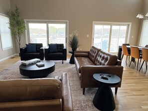 A spacious living room with a brown leather sofa, two black armchairs, a coffee table, and a dining area with chairs. - Dockside Retreat- Riverfront Near Lake Michigan (St. Joseph)