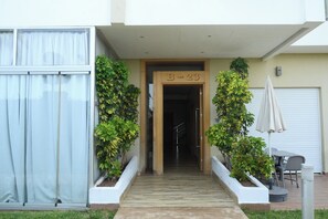 Entrada de un edificio con puerta de madera identificada con 'B-23', una escalera y macetas a ambos lados.