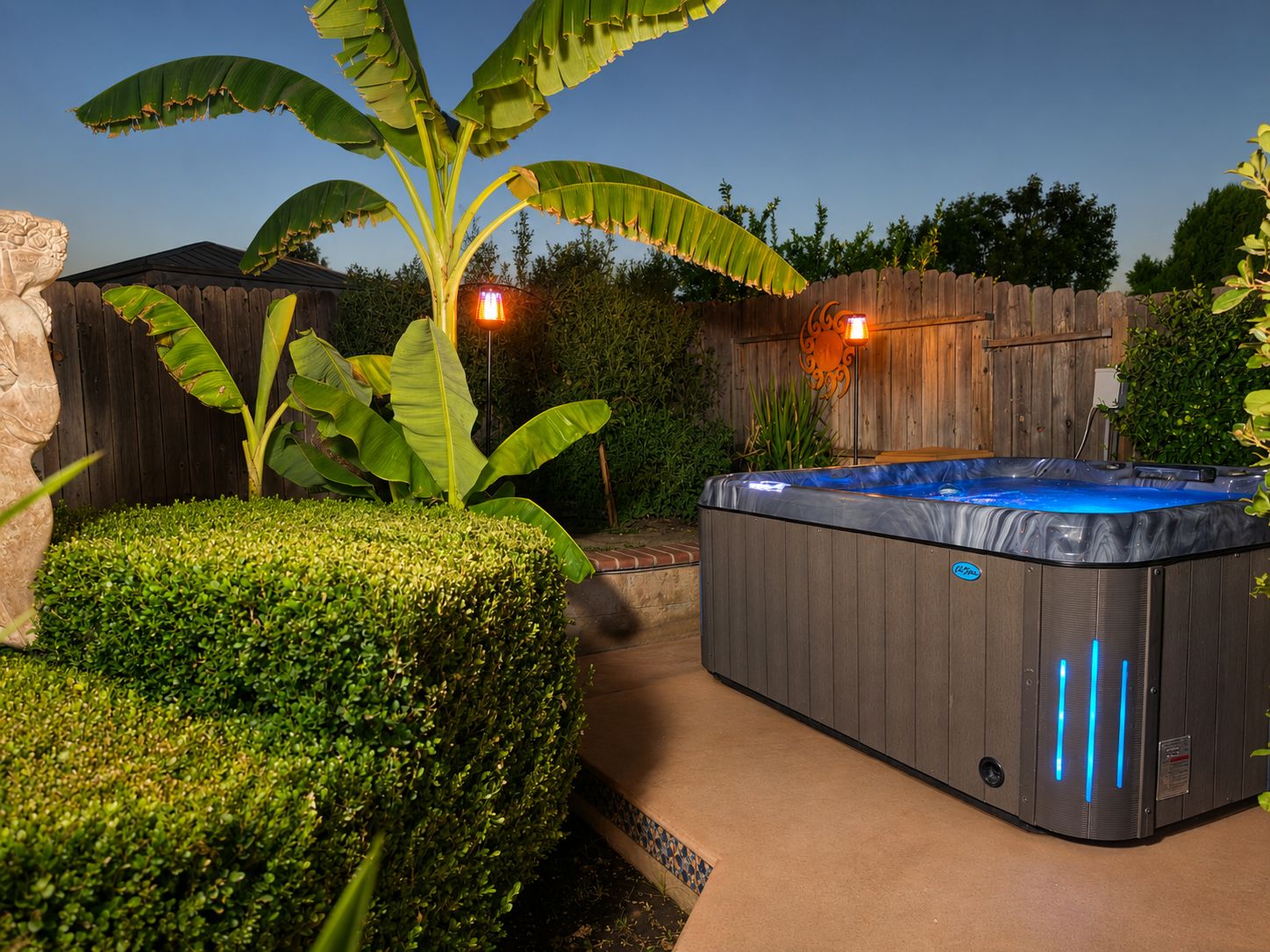 A hot tub with a wooden deck, surrounded by a wooden fence, a banana tree, and a well-maintained hedge.