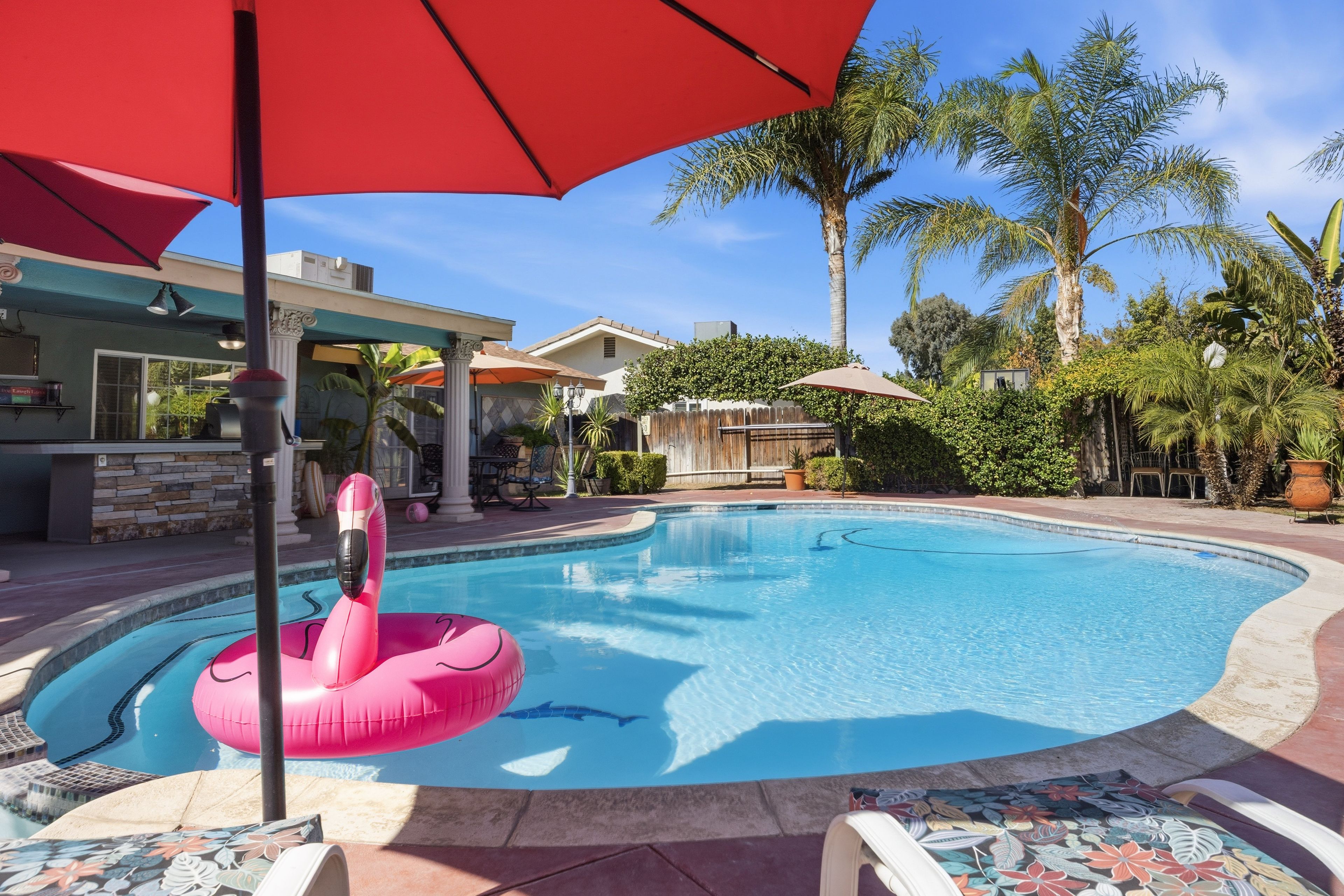 A swimming pool with a pink flamingo float, surrounded by palm trees and a patio area with umbrellas.