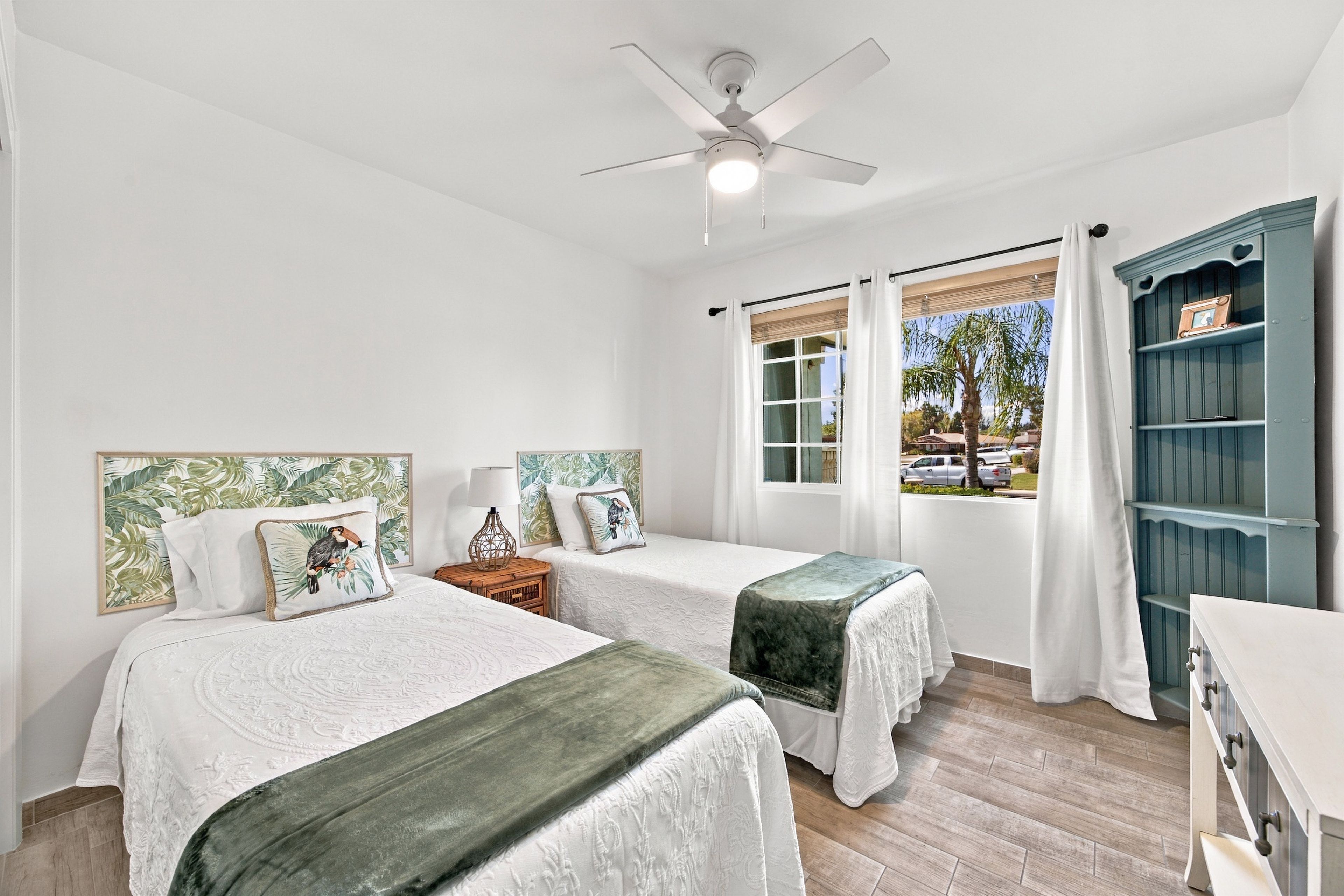 A bedroom with two beds, a ceiling fan, a window with curtains, and a wooden floor.
