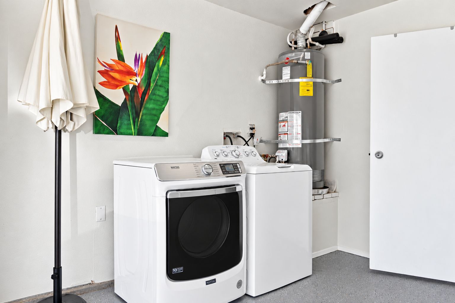 A laundry room with a front-loading washing machine and dryer, a water heater, and a painting of a flower on the wall.