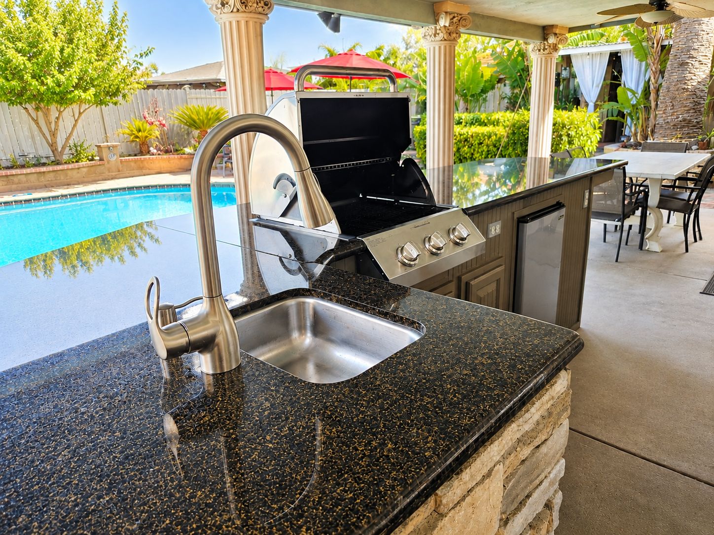 An outdoor kitchen with a sink, grill, and dining area by a pool.