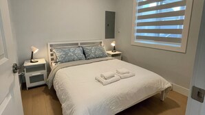 Une chambre à coucher comprenant un lit, des tables de chevet, une commode, une fenêtre avec des stores et un luminaire au plafond.