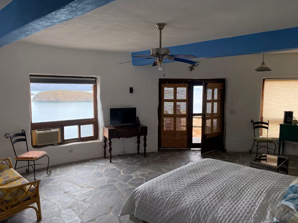 A bedroom with a bed, a television, a window with a view, a ceiling fan, and a door leading outside.. Vista a la isla .// \\. Playa Santispack