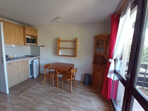 Eine kompakte Küche mit Mikrowelle, Herd und Holzschränken. Ein Esstisch mit Stühlen, ein kleiner Mülleimer und ein Fenster mit Vorhängen sind ebenfalls vorhanden.
