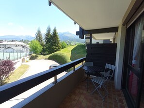 Ein Balkon mit Tisch und Stühlen, Blick auf einen Tennisplatz und eine Berglandschaft.