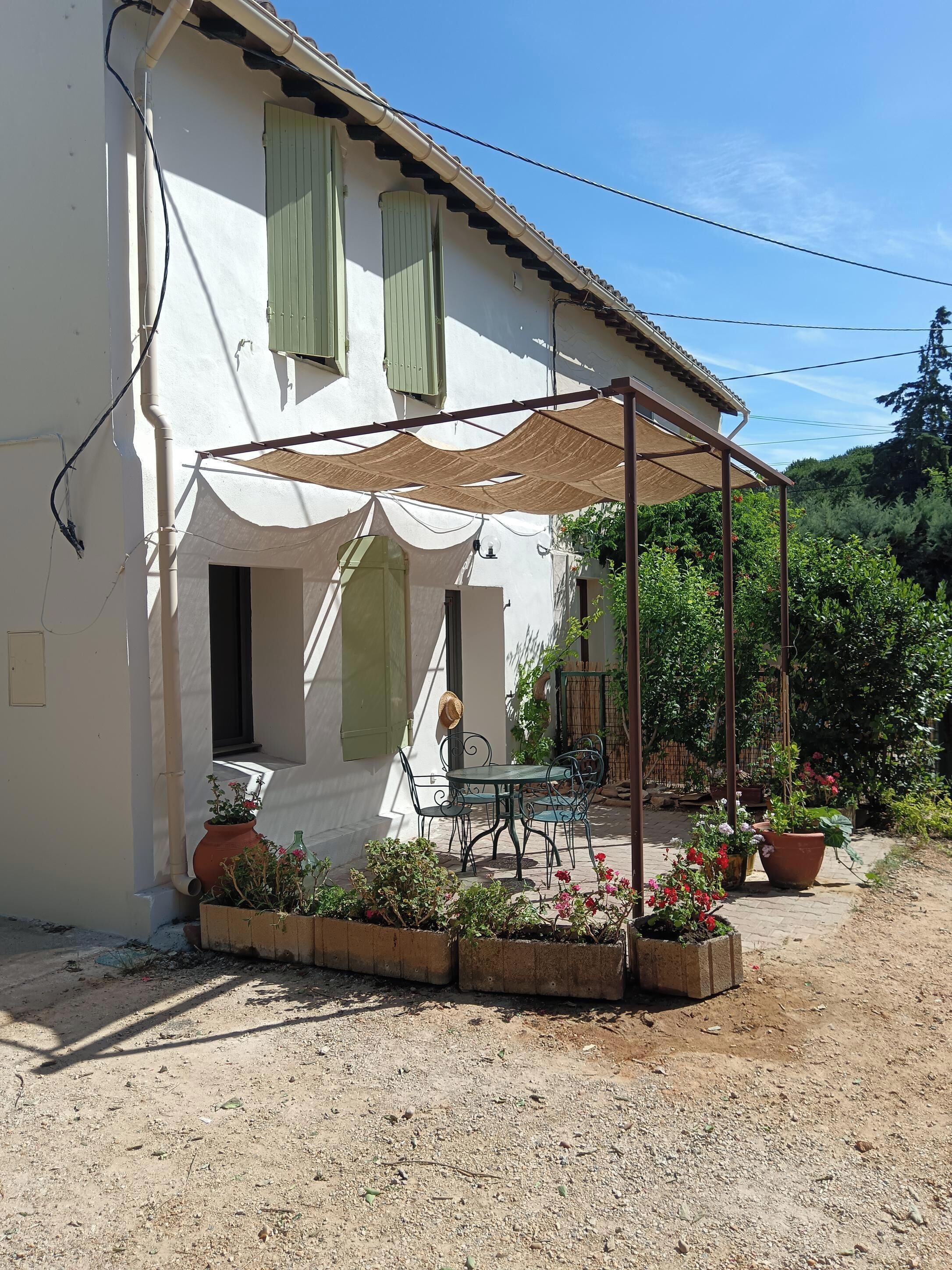 Ein weißes Gebäude mit grünen Fensterläden, eine überdachte Terrasse mit Tisch und Stühlen und Topfpflanzen.