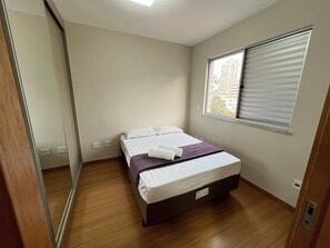 A bedroom with a bed, a window with blinds, a mirror, and a wooden floor. - LK Santa Efigênia 3 (Belo Horizonte)