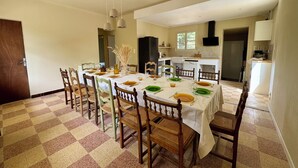 Zona de menjador amb terra de pissos, cadires de fusta, taula preparada amb plats, gots i un vas amb plantes seques.