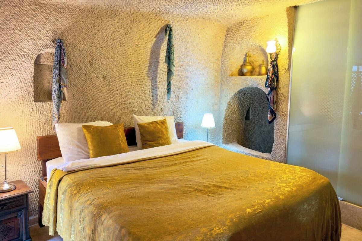 A room with a textured stone wall, a bed with a gold-colored bedspread, and a wall-mounted lamp.