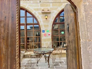 An outdoor patio with a round glass table and metal chairs, stone walls, and two large arched windows. - Gülperi Cave Otel (Nevşehir)