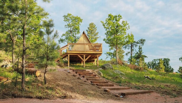 Une cabane en bois dotée d’un balcon, entourée d’arbres, avec un sentier de terre qui y mène.