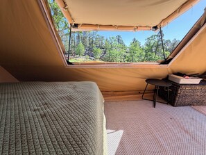 Een tent met een bed en een kleine tafel, een raam met uitzicht op bomen, en een heldere lucht.