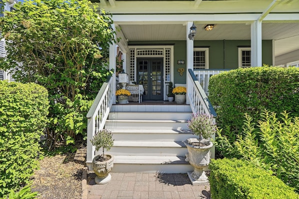 A porch with steps leading to a door, surrounded by greenery and potted plants.
