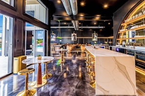 Un espace bar moderne avec des comptoirs en marbre, des tabourets de bar dorés et une vue sur l’extérieur.