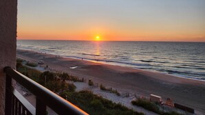 En vy över stranden med solen som går ner över havet, en sandstrand och några få personer som promenerar.