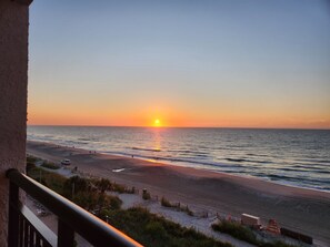 Une vue sur la plage, avec le soleil se couchant sur l’océan, une plage de sable et quelques personnes qui se promènent.