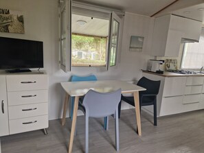 A modern kitchen with a dining area, a TV on a cabinet, and a window with a view of greenery. - Mobile home in leisure residence (Saint-Gilles-Croix-de-Vie)