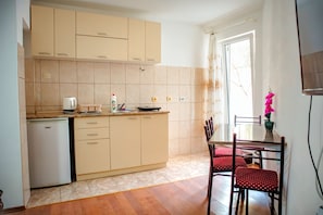A kitchen with beige cabinets, a refrigerator, a dining table with chairs, and a window with a curtain. - Apartmani Grbovic (Čanj)