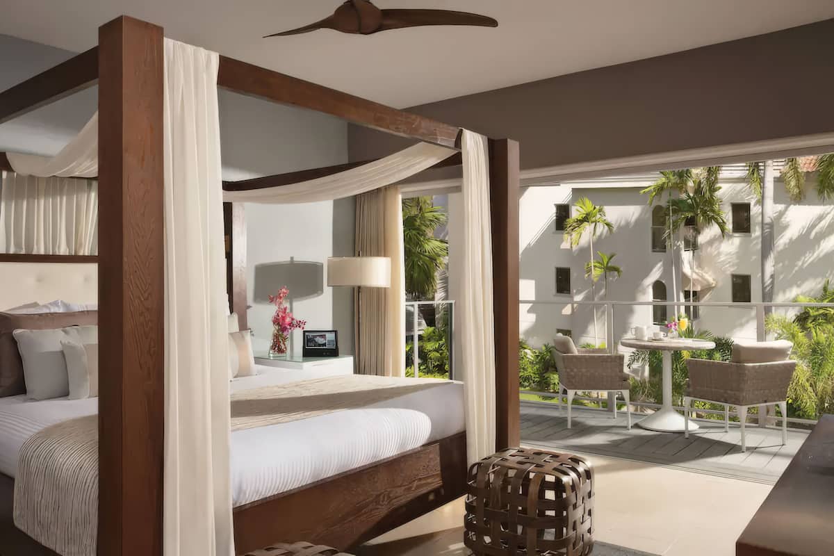 Uma cama com dossel e cortina, uma mesa de cabeceira com abajur, e vista para uma varanda com mobiliário.