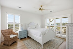 A bedroom with a bed, a nightstand, a wicker chair, and a balcony with a railing. - Steps from Beach with Ocean Views and Fresh Updates (Oak Island)