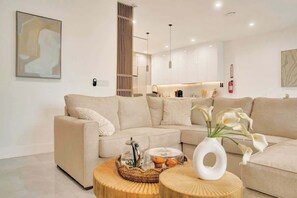 Ein modernes Wohnzimmer mit einem beigefarbenen Ecksofa, einem runden Couchtisch mit Blumen und einer Küche im Hintergrund.