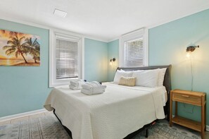 Quarto com cama, duas janelas com persianas, pintura de palmeiras, mesa de cabeceira com abajur e tapete estampado.
