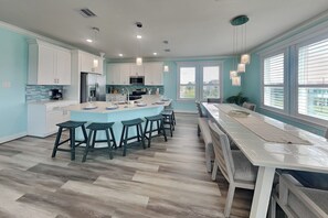 Une cuisine moderne avec des meubles blancs, un îlot central doté de tabourets, un coin repas avec une longue table et de grandes fenêtres.