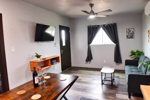 A living room with a wooden dining table, a sofa, a TV mounted on the wall, a ceiling fan, and a window with curtains. - 22 Eleven Claystone cozy cabin BBQ grill, Firepit, Pergola (Devine)
