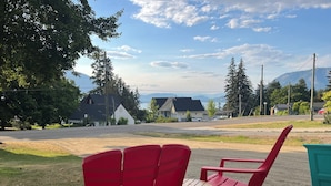 A view of a residential street with houses, a clear sky, and a grassy area with red chairs and a small table. -        Salmon Arm Comfortable 2 Bedroom in town, 5 minute drive to the beach! (Salmon Arm)