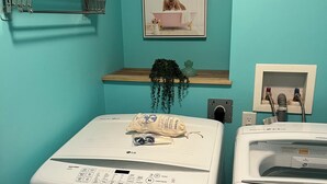 A laundry room with two white front-loading washing machines, a shelf with a plant, and a framed picture on the wall. -        Salmon Arm Comfortable 2 Bedroom in town, 5 minute drive to the beach! (Salmon Arm)