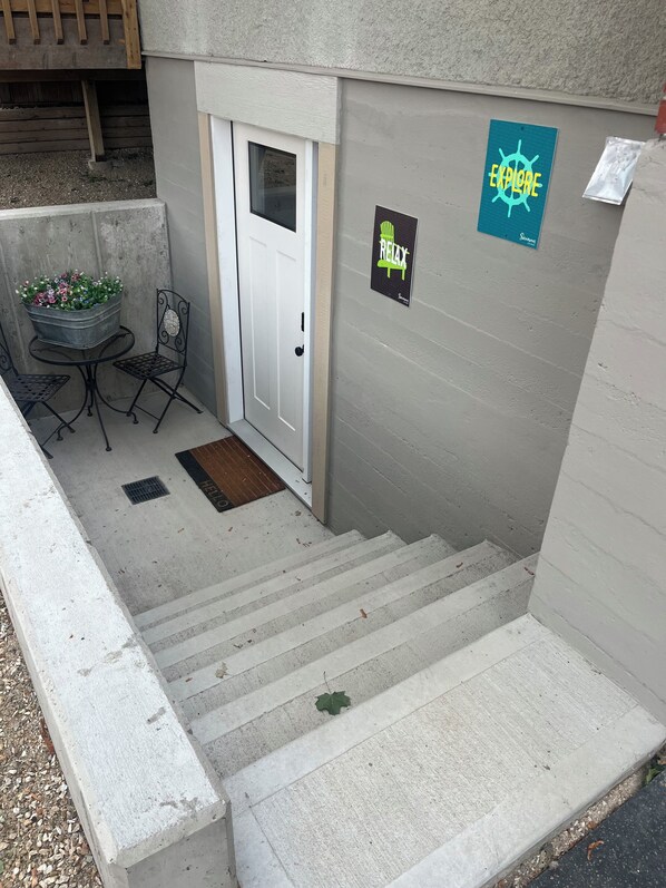 Unos escalones de concreto conducen a una puerta, un pequeño espacio exterior con sillas de metal, y dos carteles en la pared.