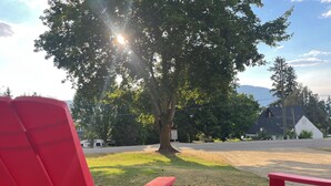 A red chair and a round wooden table in a grassy area with a tree and a house in the background. -        Salmon Arm Comfortable 2 Bedroom in town, 5 minute drive to the beach! (Salmon Arm)