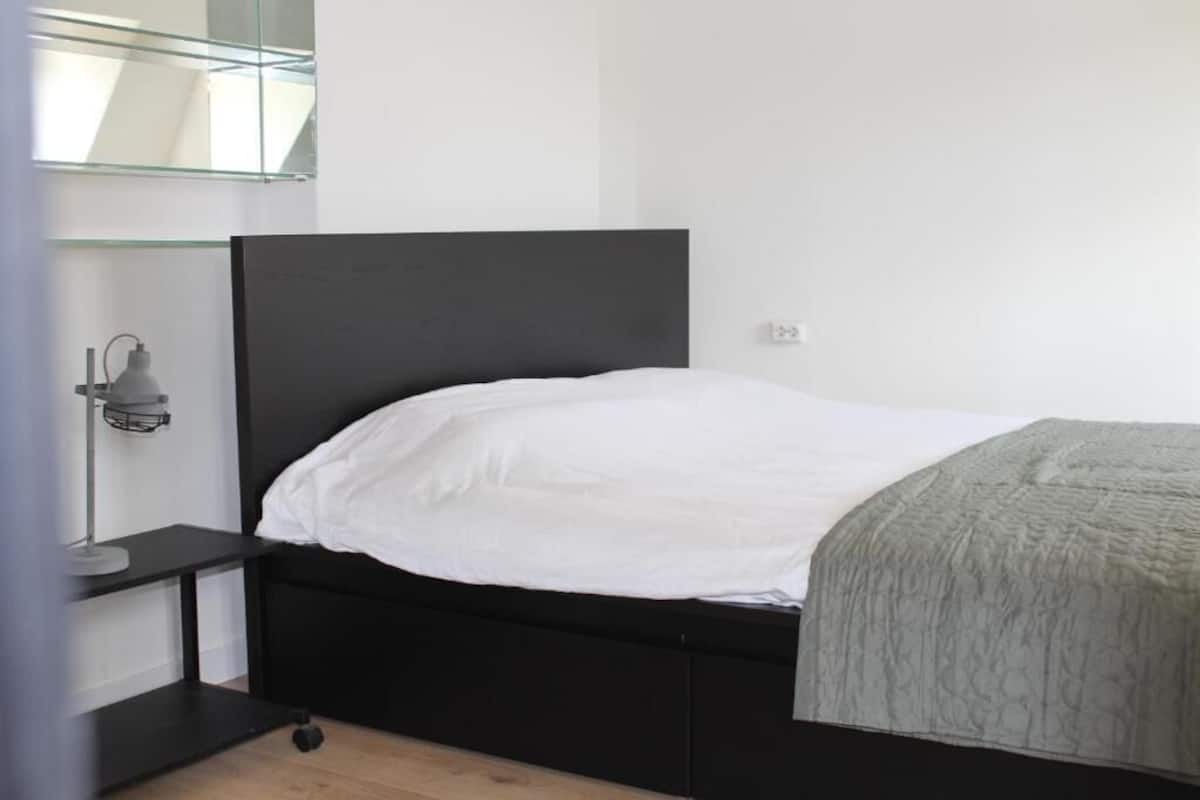 A modern bedroom with a black bed frame, white bedding, a bedside table, and a wall-mounted lamp.