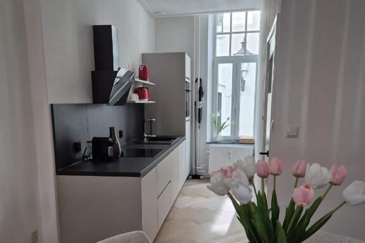 A modern kitchen with white cabinets, a black countertop, and a vase of pink tulips on a round table.