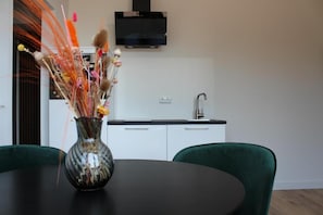 A dining area with a round table, two green chairs, a vase with dried flowers, a black TV mounted on the wall, and a kitchen area with white cabinets and a sink.