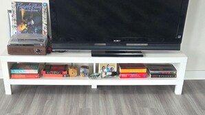 Uma televisão de tela plana em um suporte branco, com uma prateleira inferior que acomoda livros e um aparelho de CD.