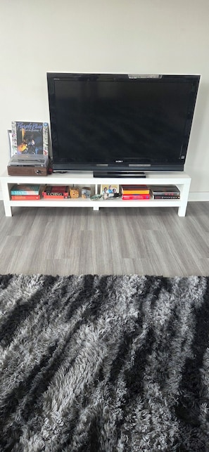 A flat-screen TV on a white stand with a shelf below holding books and a CD player. - 15 min from World Cup stadium! Soothing 2-bd room apt w/AC in Grand Prairie (Grand Prairie)