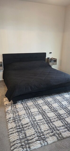 A bedroom with a black bedspread, a patterned rug, and a nightstand with a lamp. - 15 min from World Cup stadium! Soothing 2-bd room apt w/AC in Grand Prairie (Grand Prairie)