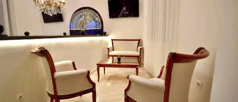 Una habitación con barra, dos sillones tapizados, una mesita, una araña y un amplio ventanal de arco, además de un televisor de pantalla plana montado en la pared.