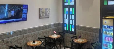 Un restaurante con mesas redondas, sillas negras, una araña de cristal, un televisor y una máquina expendedora.