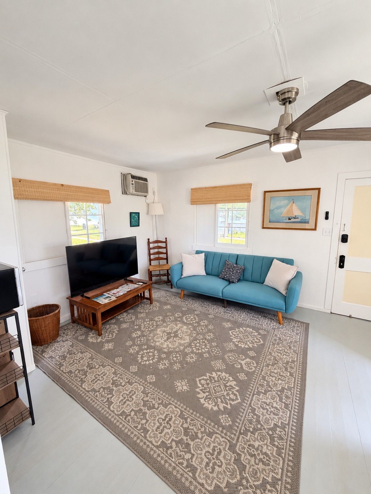 A living room with a flat-screen TV, a blue sofa, a patterned rug, a ceiling fan, and a window with a view.