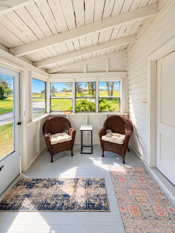 A sunroom with white walls, two wicker chairs, a small table, and a view of a green landscape through large windows.