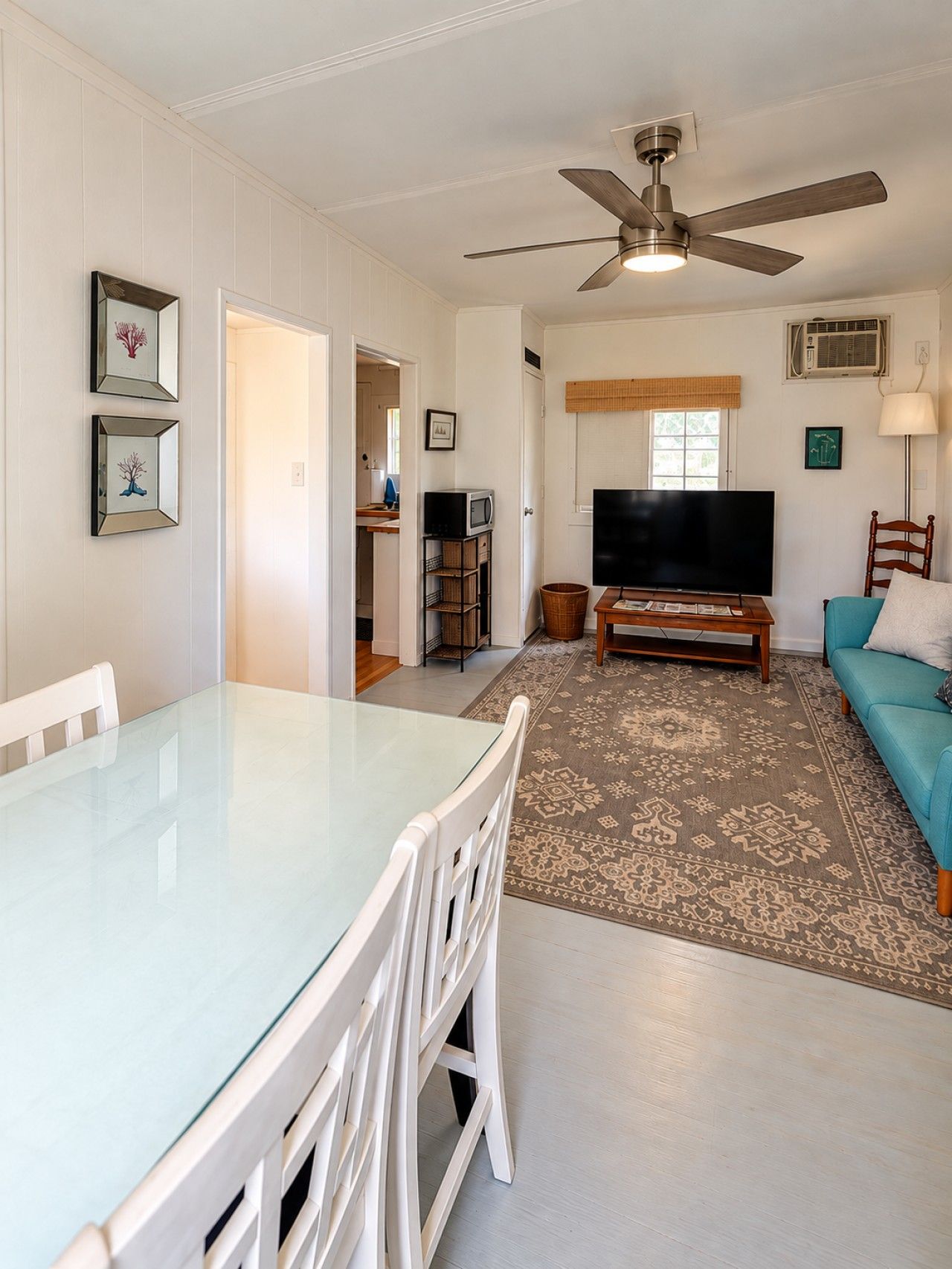 A living room with a glass-top dining table, white chairs, a sofa, a television, a ceiling fan, and a patterned rug.